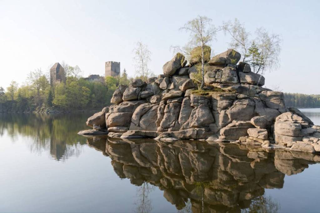 Ruhiges Wasser am Stausee Ottenstein spiegelt eine große, von Birken bewachsene Felsformation wider. Im Hintergrund ragt die Burgruine Lichtenfels malerisch aus den grünen Baumwipfeln.