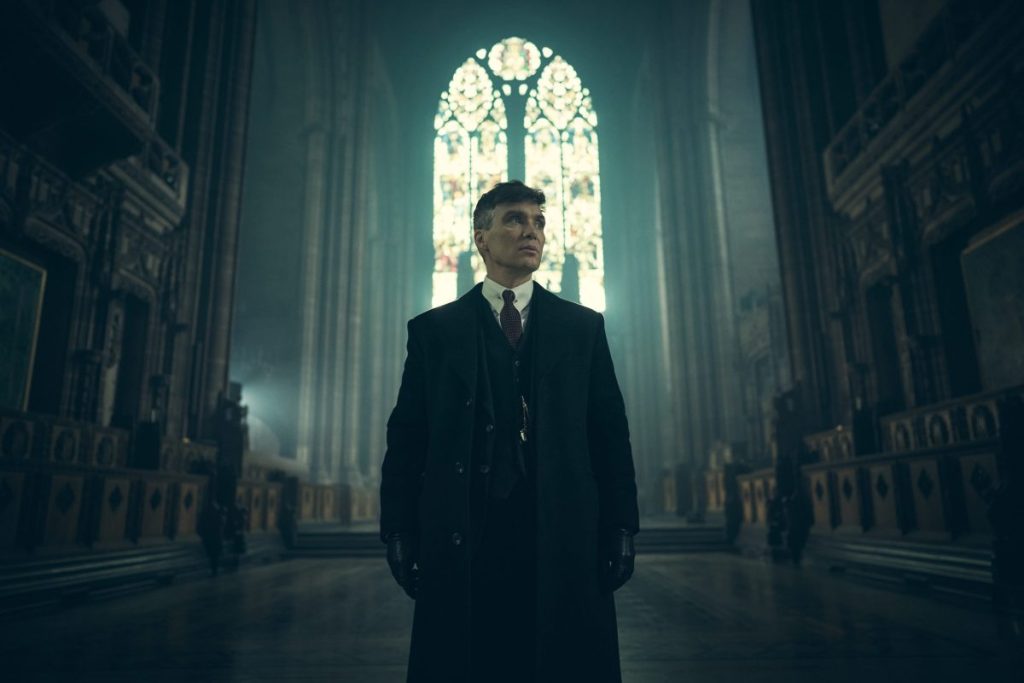 Tommy Shelby, vestido con un traje oscuro y abrigo largo, en el interior de una iglesia con grandes vitrales de fondo, en una escena de Peaky Blinders.